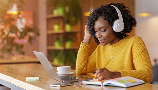 带着无线耳机的微笑黑人女孩在网上学习，在咖啡馆用笔记本电脑，做笔记